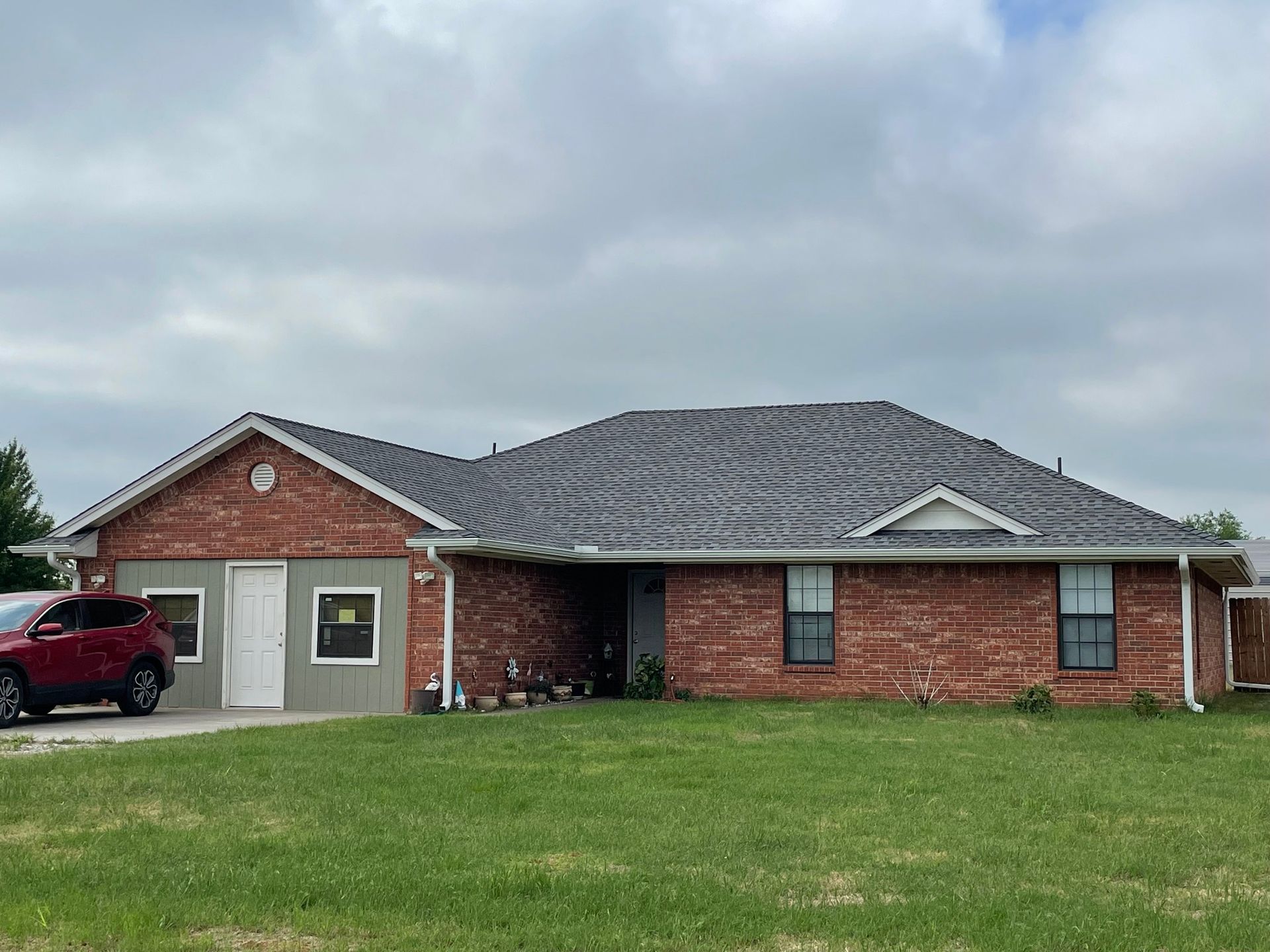 A red car is parked in front of a brick house with a gray roof.