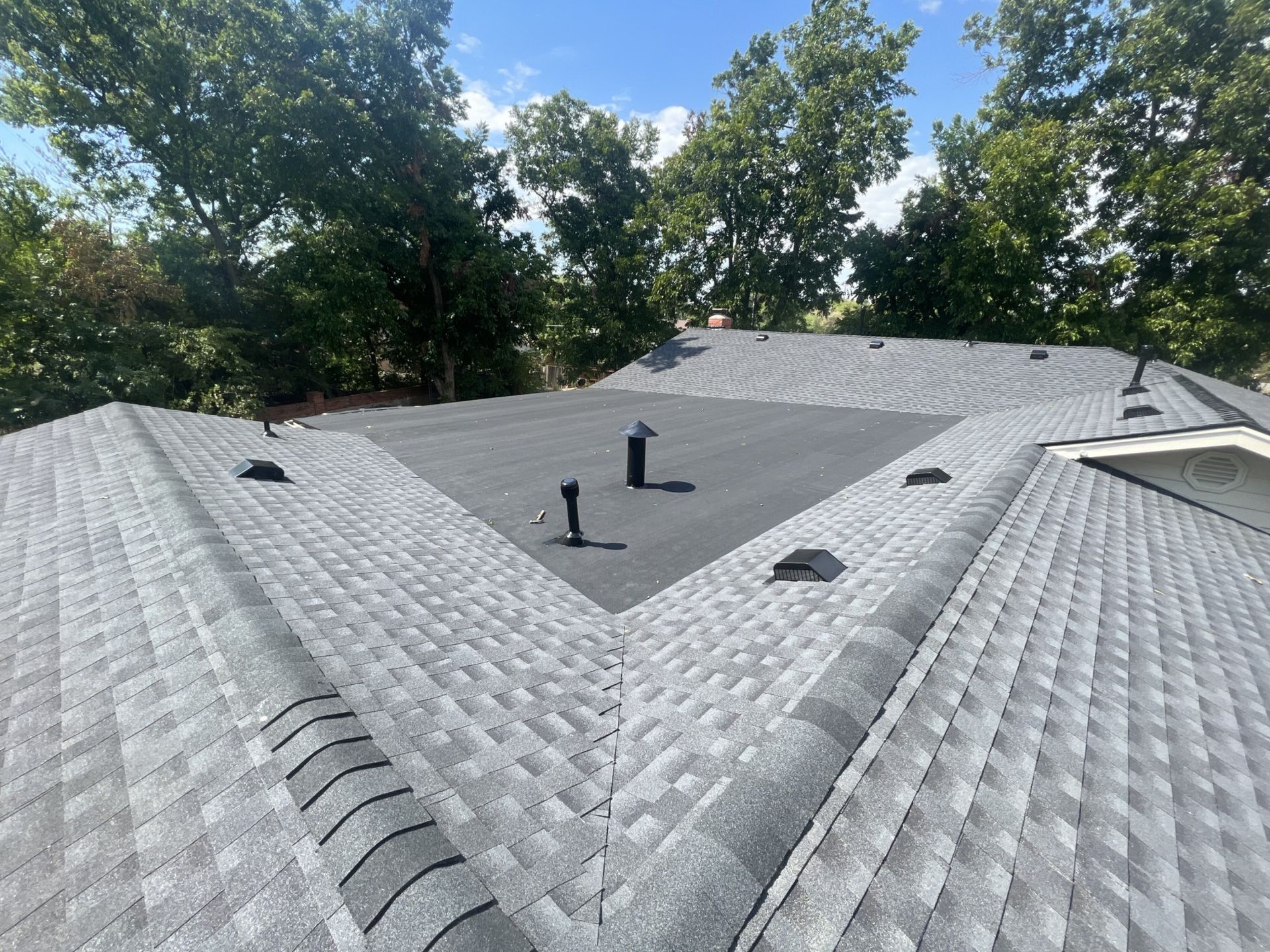 Grey shingle roof with black vents and flat central area, trees in the background under a bright sky.