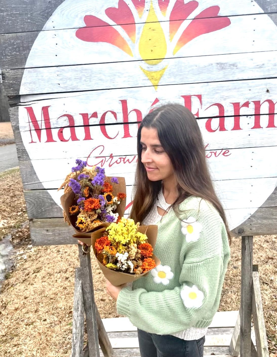 A woman is holding two bouquets of flowers in front of a march farm sign.