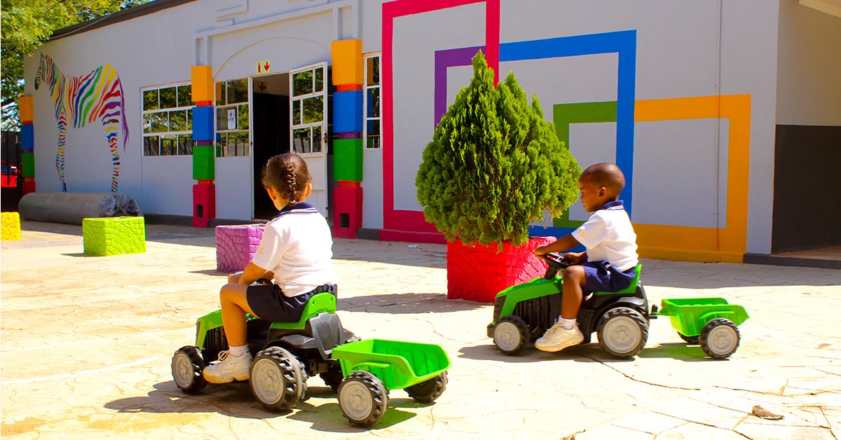 Two children are riding toy tractors in front of a building.
