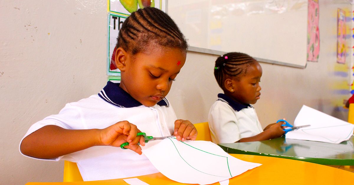 Two young girls are sitting at a table cutting paper with scissors.