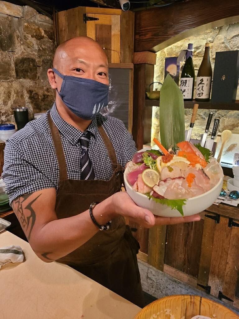 Man in a mask and apron presenting a colorful sashimi bowl in a rustic restaurant