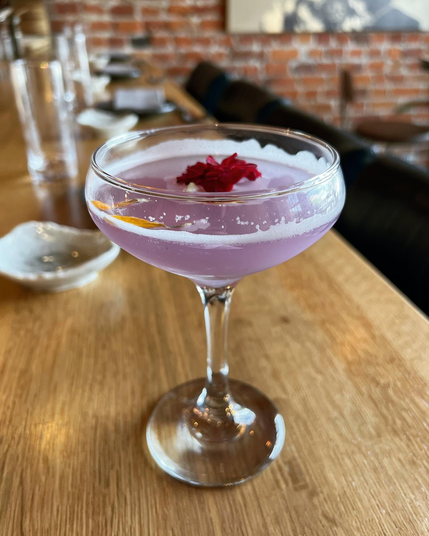 Lavender cocktail in a coupe glass on a wooden bar, garnished with red petals