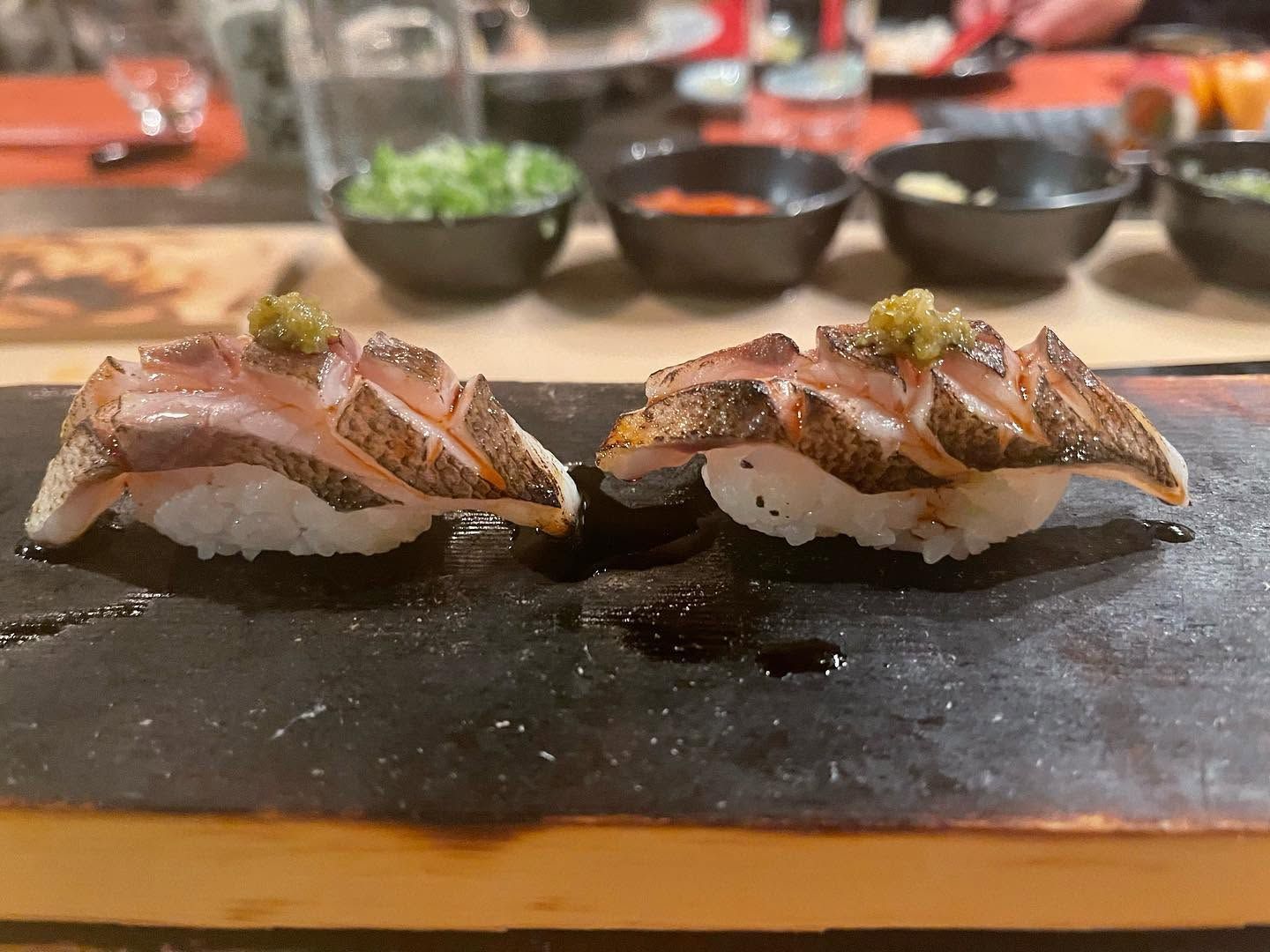 Two pieces of seared sushi on a black stone plate with small dipping bowls in the background