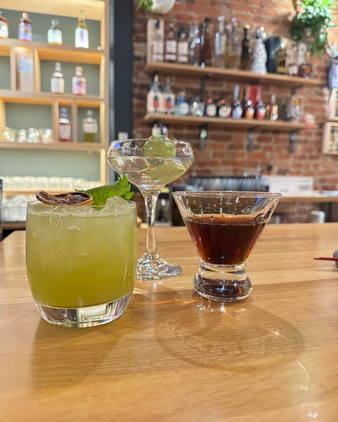 Three cocktails on a bar table, including a green margarita and a martini, with a warm brick bar background