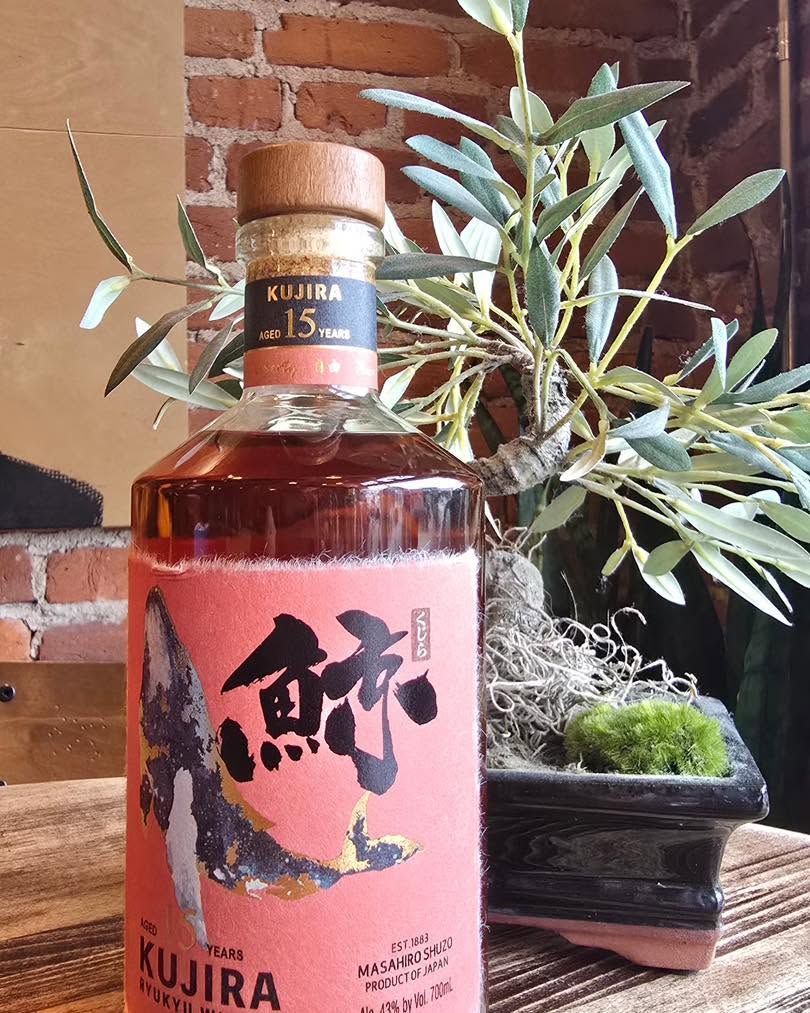 Kujira whiskey bottle with pink label beside a bonsai plant on a wooden table against a brick wall.