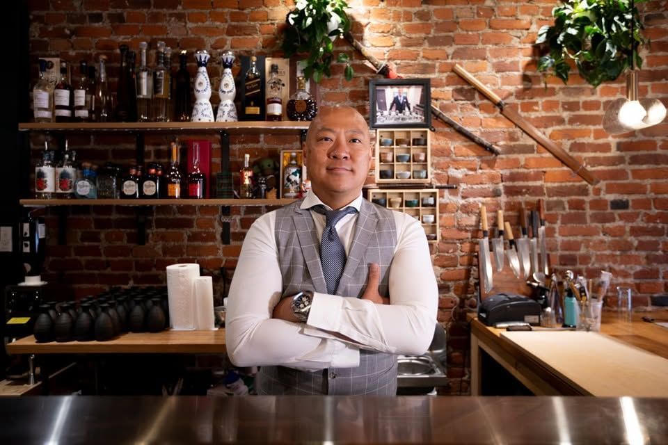Bar owner stands behind a bar in a brick-walled cafe, arms crossed, with bottles and plants on shelves.