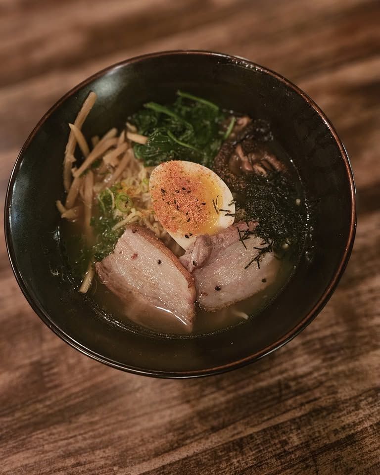 Bowl of ramen with sliced pork, soft-boiled egg, seaweed, and greens on a wooden table