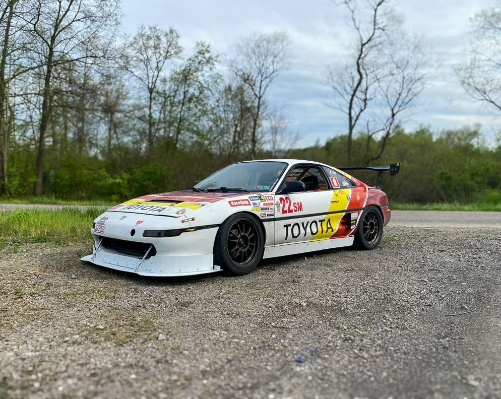White and yellow Toyota MR2 race car on a gravel road.  Black wheels, large spoiler, and racing livery.