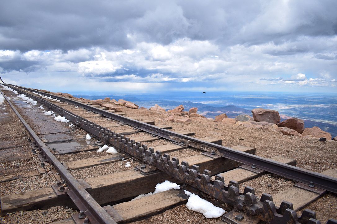 Railway tracks on a mountain summit, with snow patches and distant landscape under a cloudy sky.