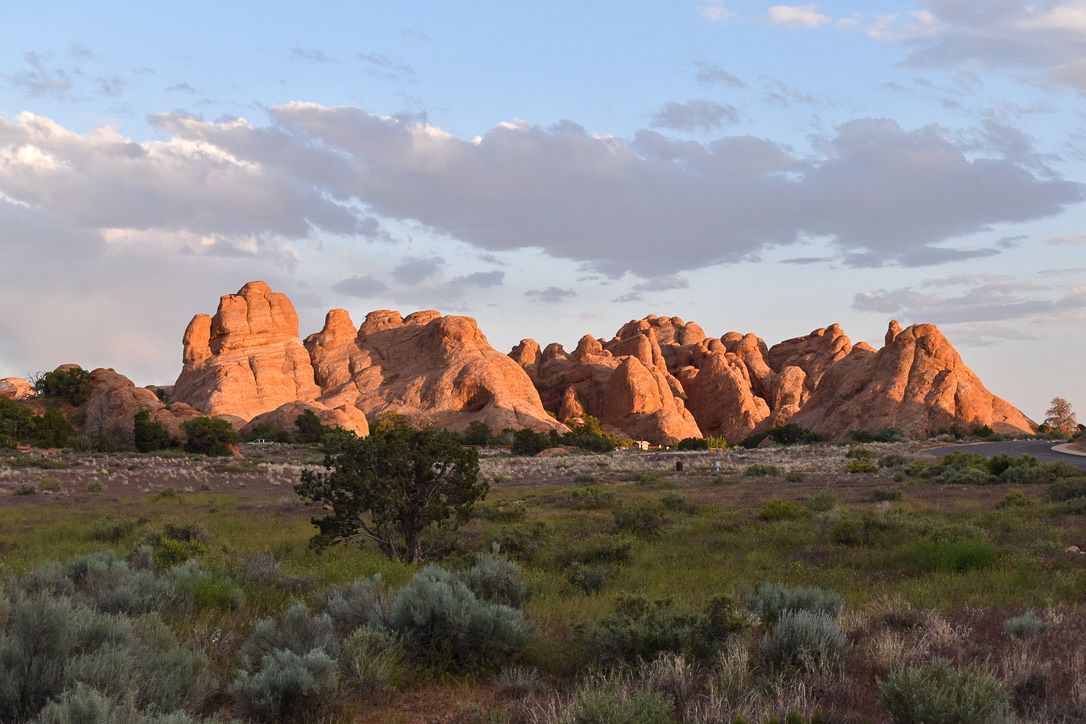 Red rock formations against a blue sky, lit by sunlight, with green vegetation in the foreground.