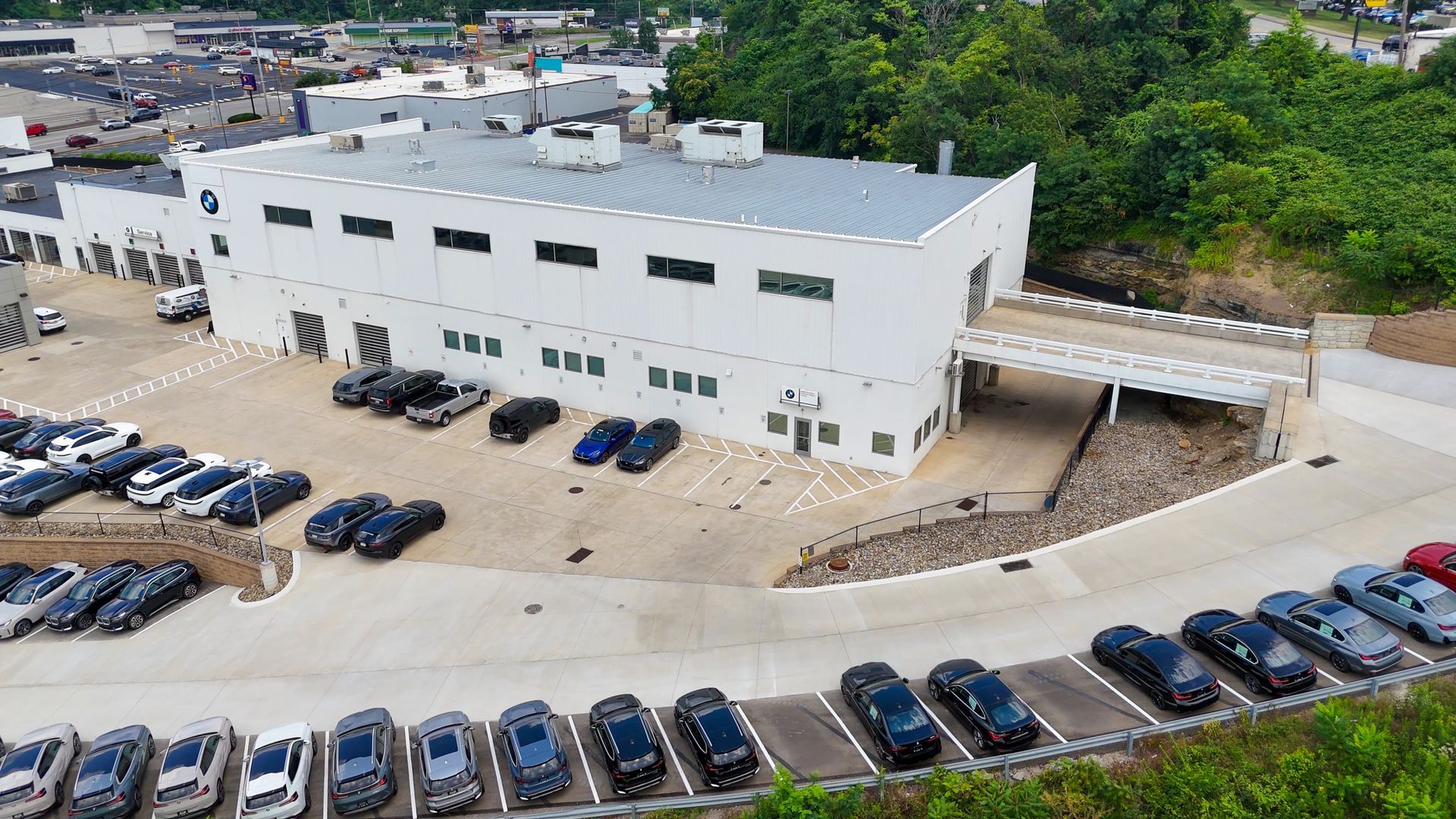 White building with cars parked in front. Trees on the right and a parking lot in the background.