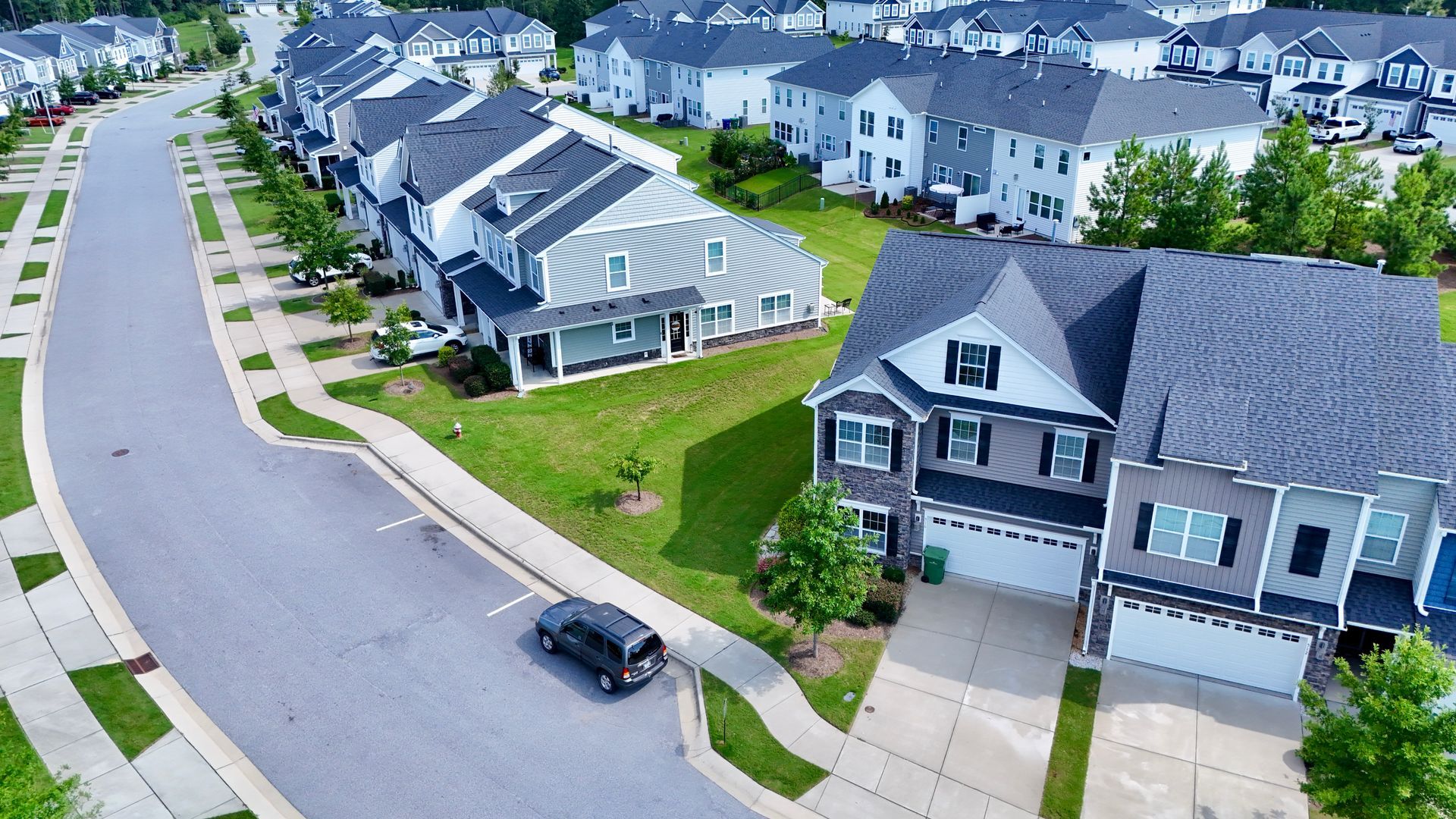 Aerial view of a residential neighborhood with gray houses, paved streets, and a dark SUV parked.