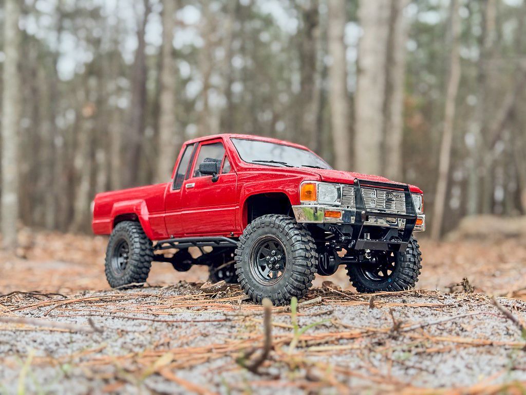 Red RC truck on a forest floor with trees in the background.