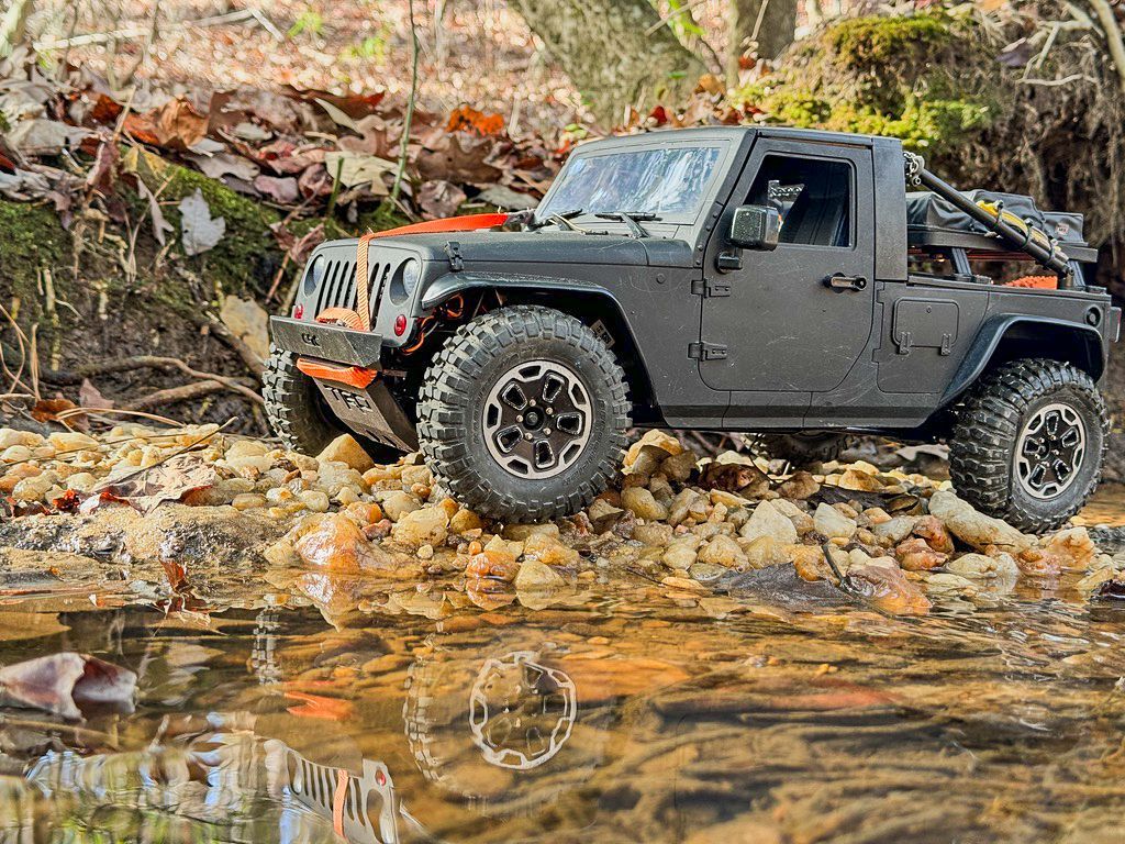 Black RC Jeep navigating a rocky creek bed, its reflection in the water.