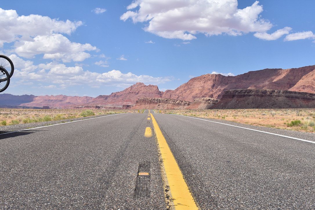Road through a desert landscape with red rock formations under a blue sky with clouds.
