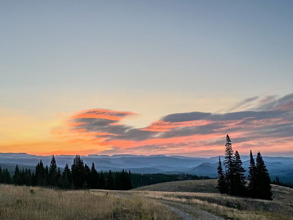 Sunrise over rolling hills and a forest, with colorful sky.