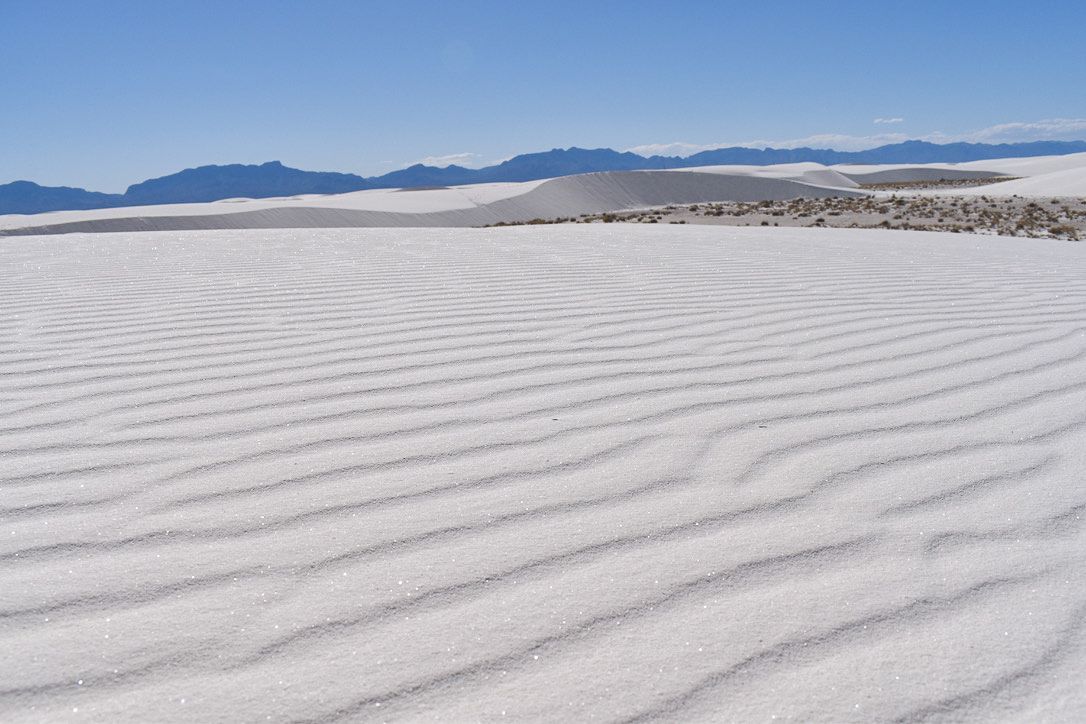White sand dunes with textured ridges, distant mountains under a clear blue sky.