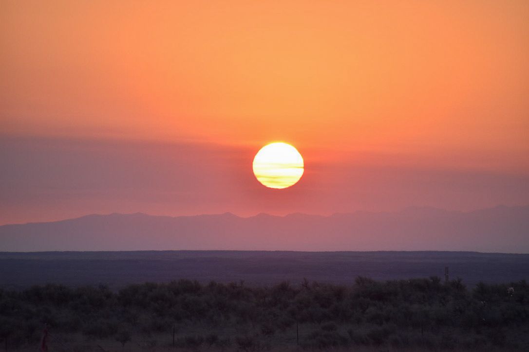 Sunset over a distant mountain range; orange sky, sun, and silhouetted desert landscape.