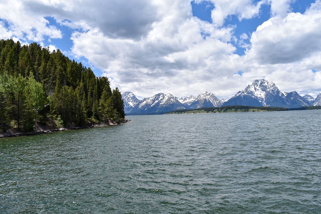 Lake with evergreen forest on one side, snow-capped mountains in the distance, under a cloudy sky.