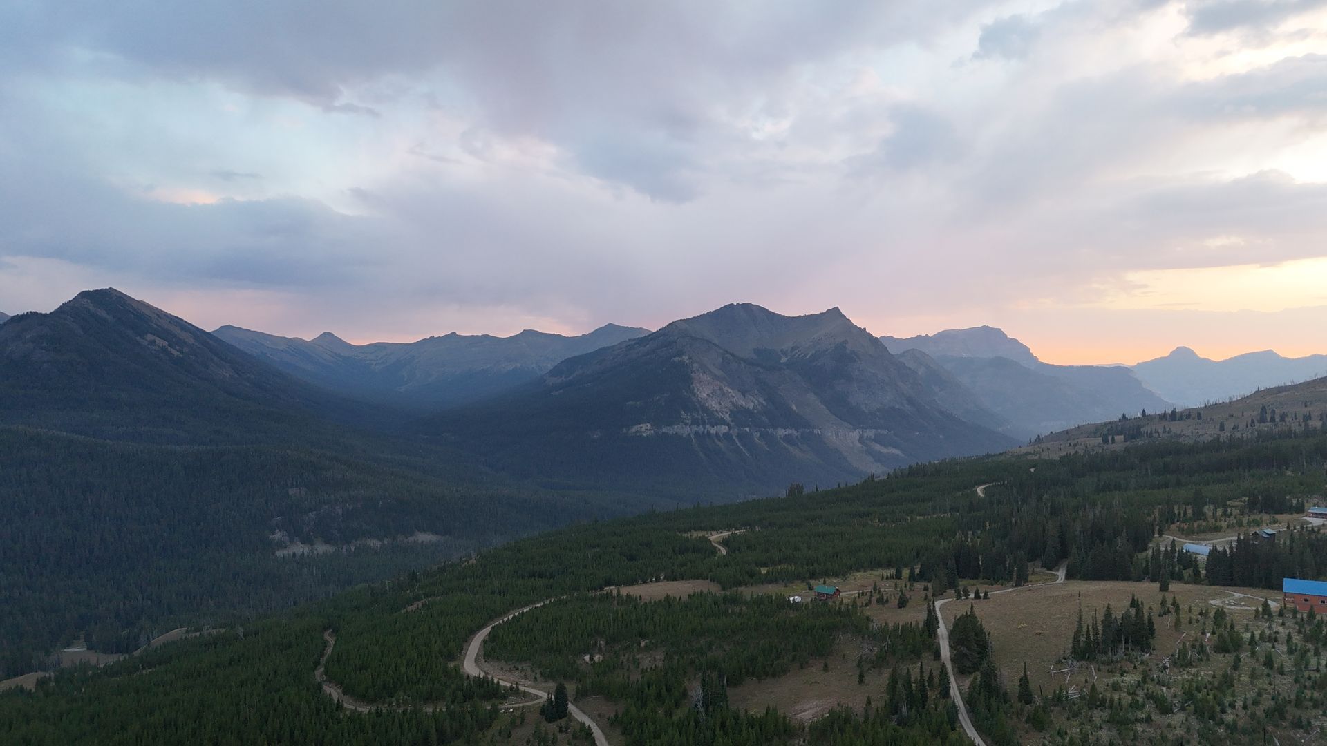 Mountainous landscape with cloudy sky; dark green forests and patches of gold-colored grass in valley below.