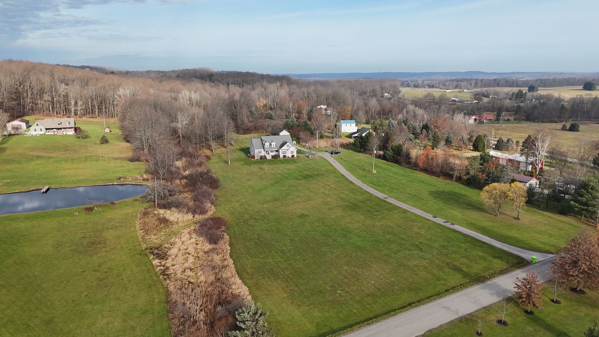 Aerial view of a house on a grassy hill with a pond, surrounded by trees and fields on a sunny day.