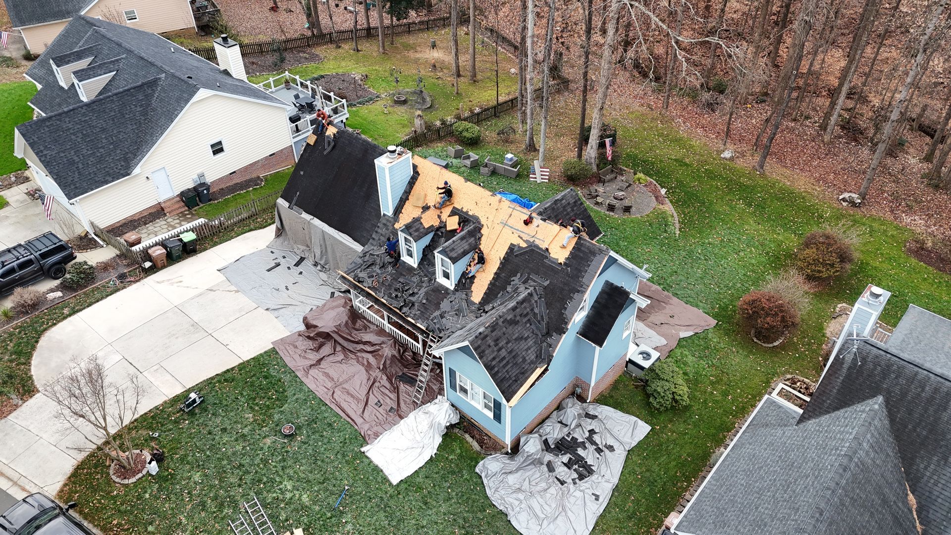 Aerial view of a blue house with roof damage, surrounded by green grass, tarps, and a driveway.