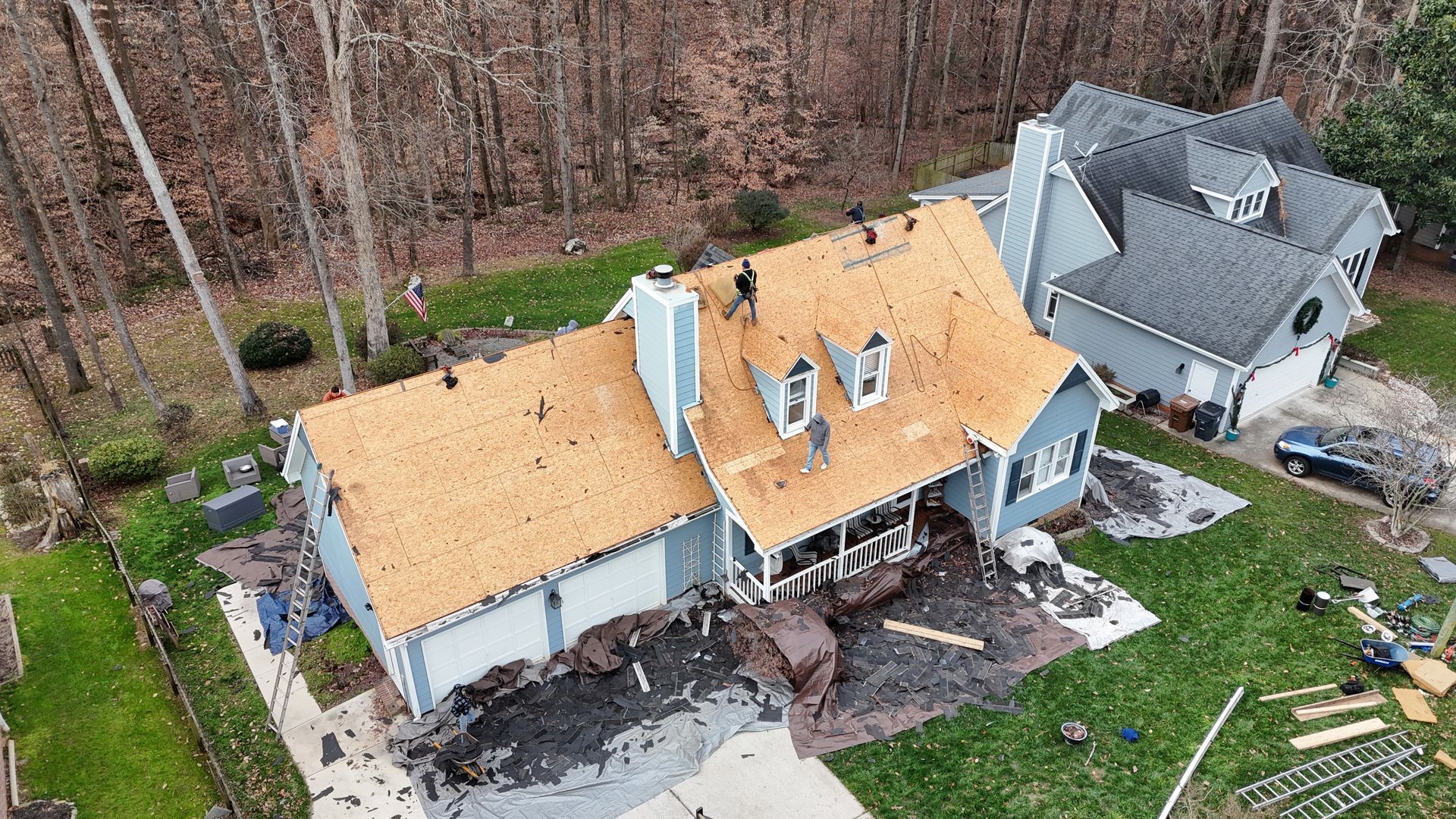 Roofers replacing a blue house's roof, debris scattered on the ground, adjacent to a gray house.