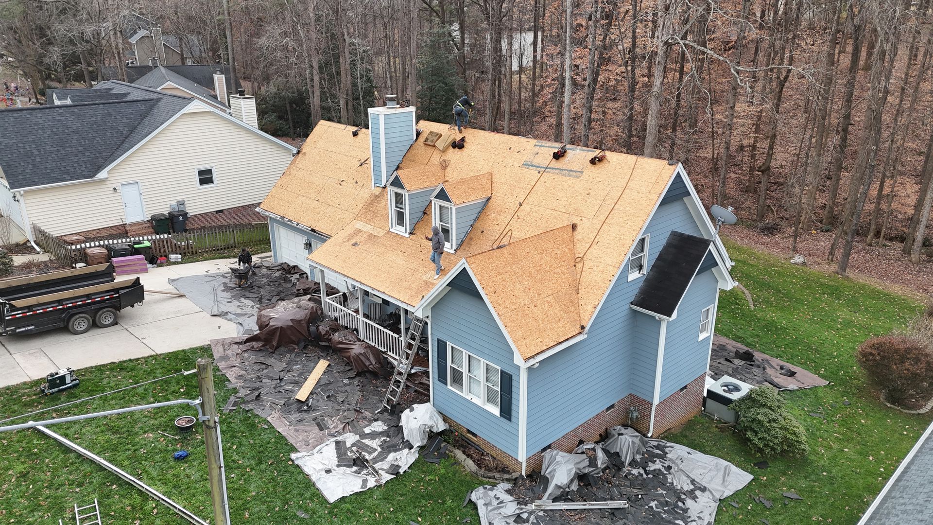 House with a new roof under construction; debris on the ground; blue siding; leafless trees in the background.