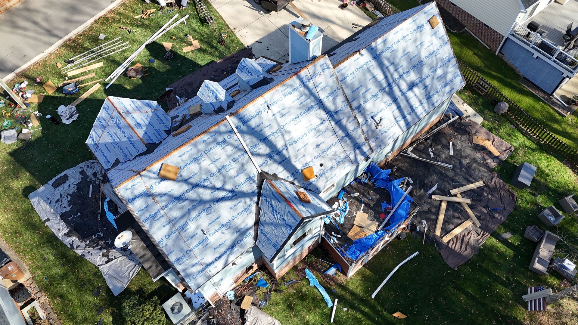 Aerial view of a house undergoing roof replacement; roof exposed, materials scattered on the lawn.