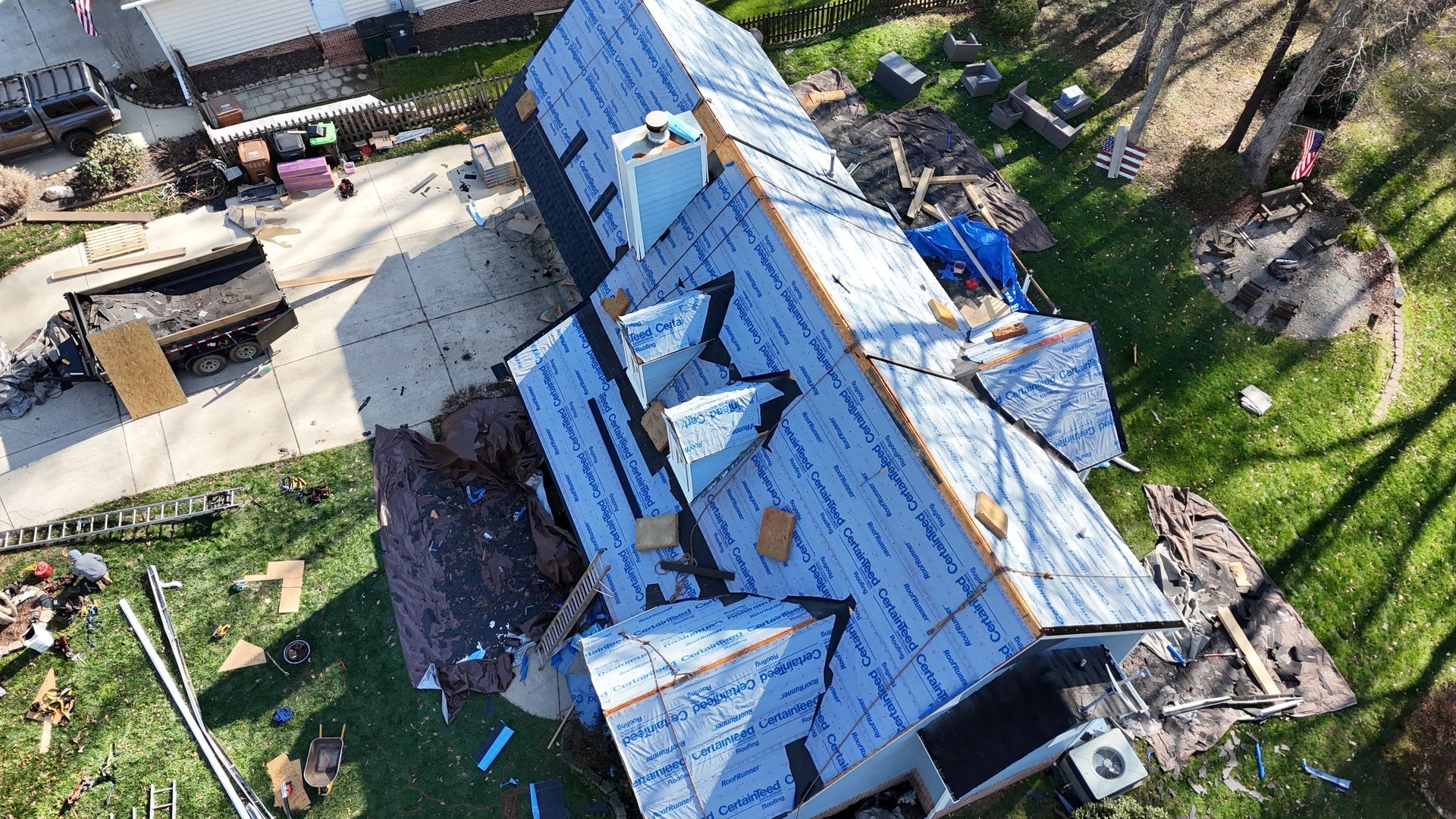 Aerial view of a house with a partially torn-off roof, construction materials, and a surrounding yard.