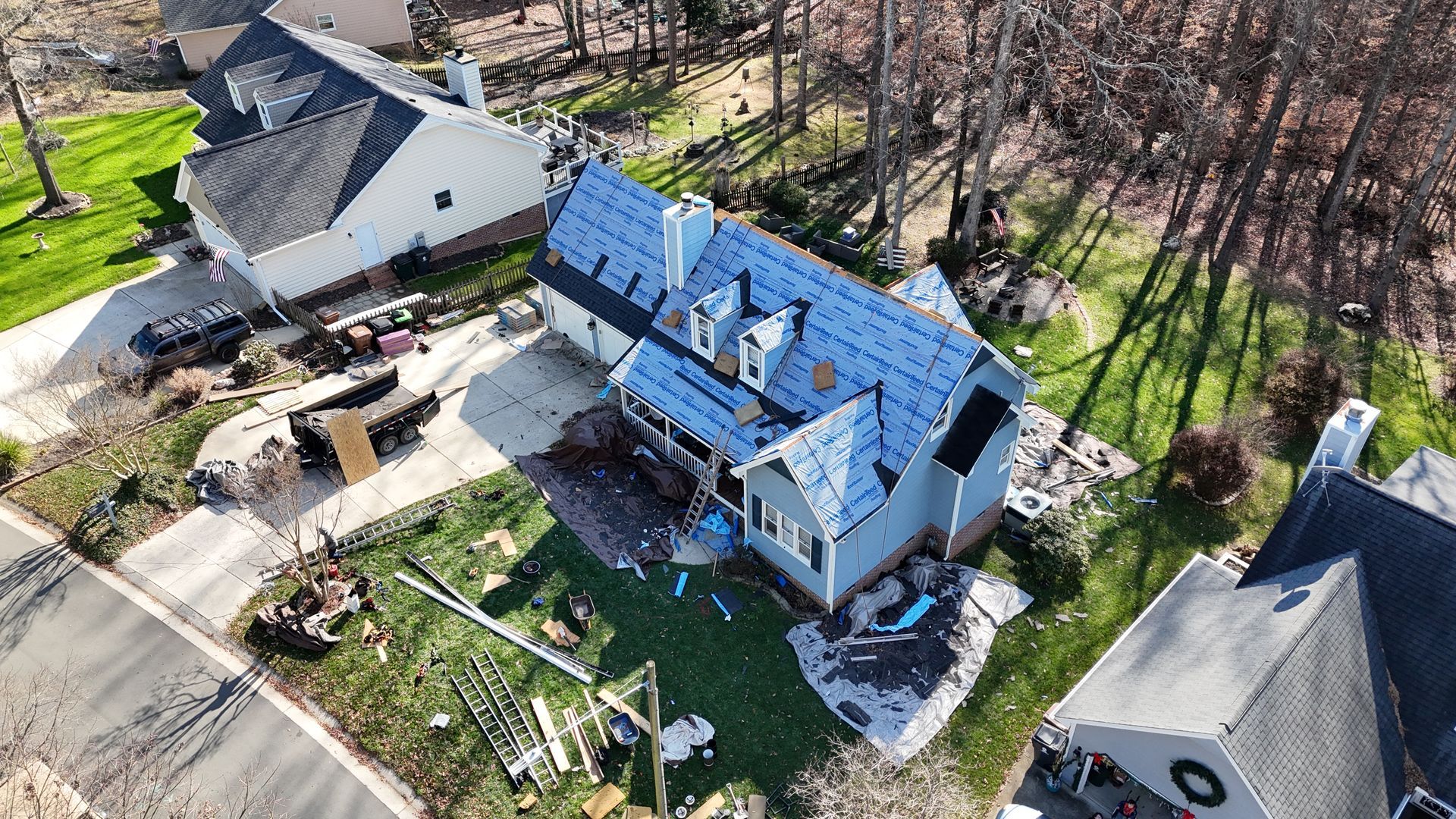 Aerial view of a house with significant roof damage, surrounded by debris on a sunny day.