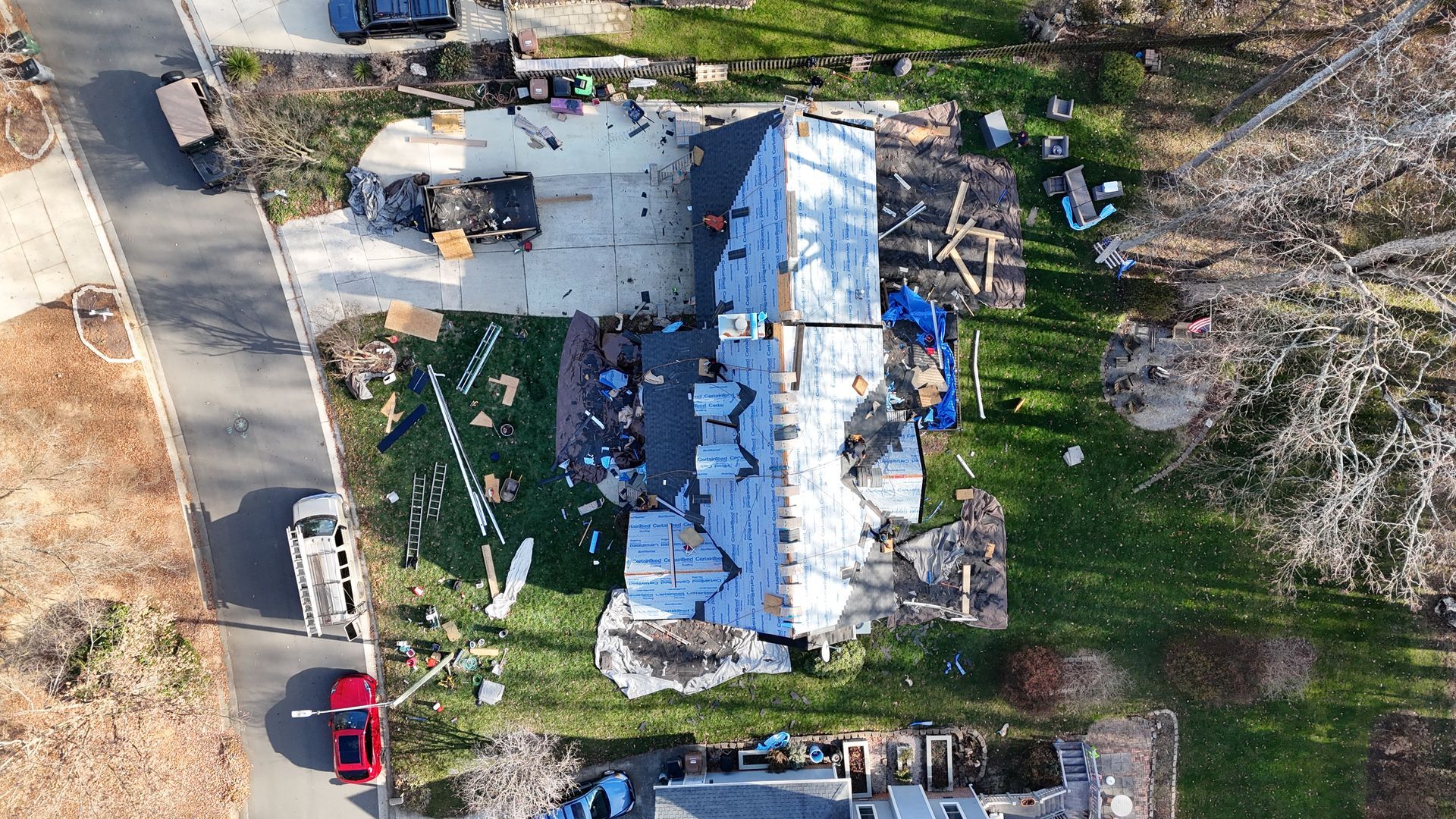Aerial view of a house with significant roof damage, debris scattered on the yard, and vehicles parked nearby.