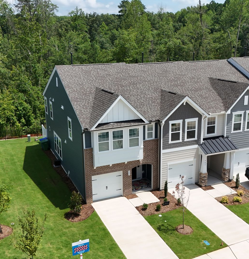 Townhomes with green siding, brick accents, and white garage doors against a background of green trees under a blue sky.