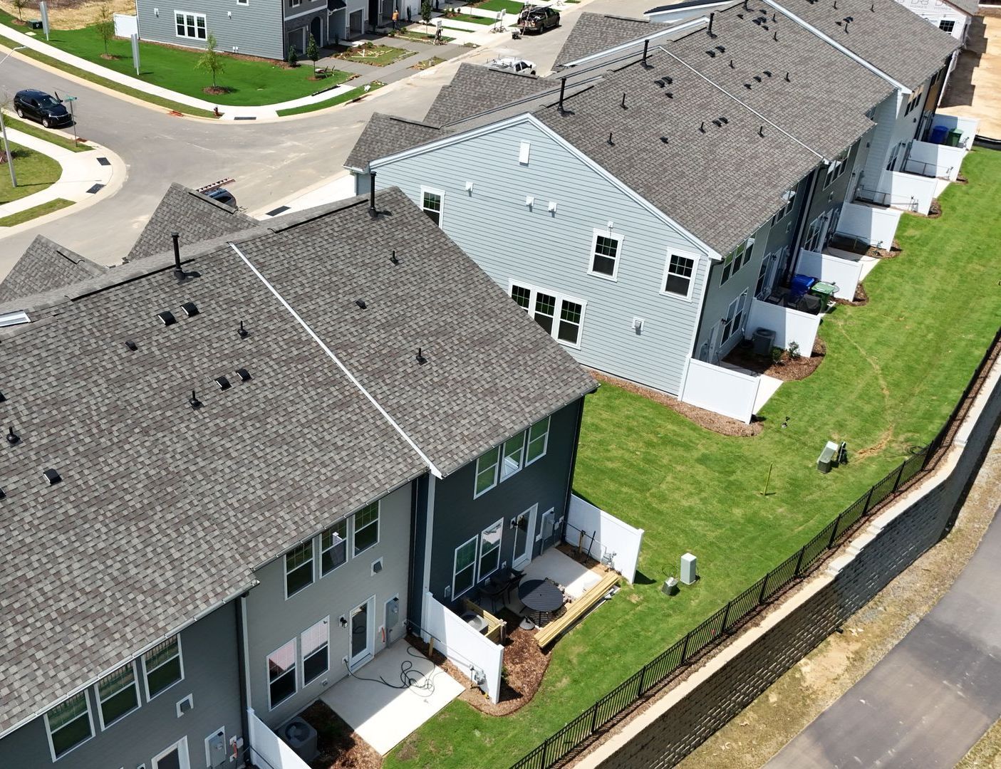 Row of gray townhouses with green grass, a black fence, and a street in the background.