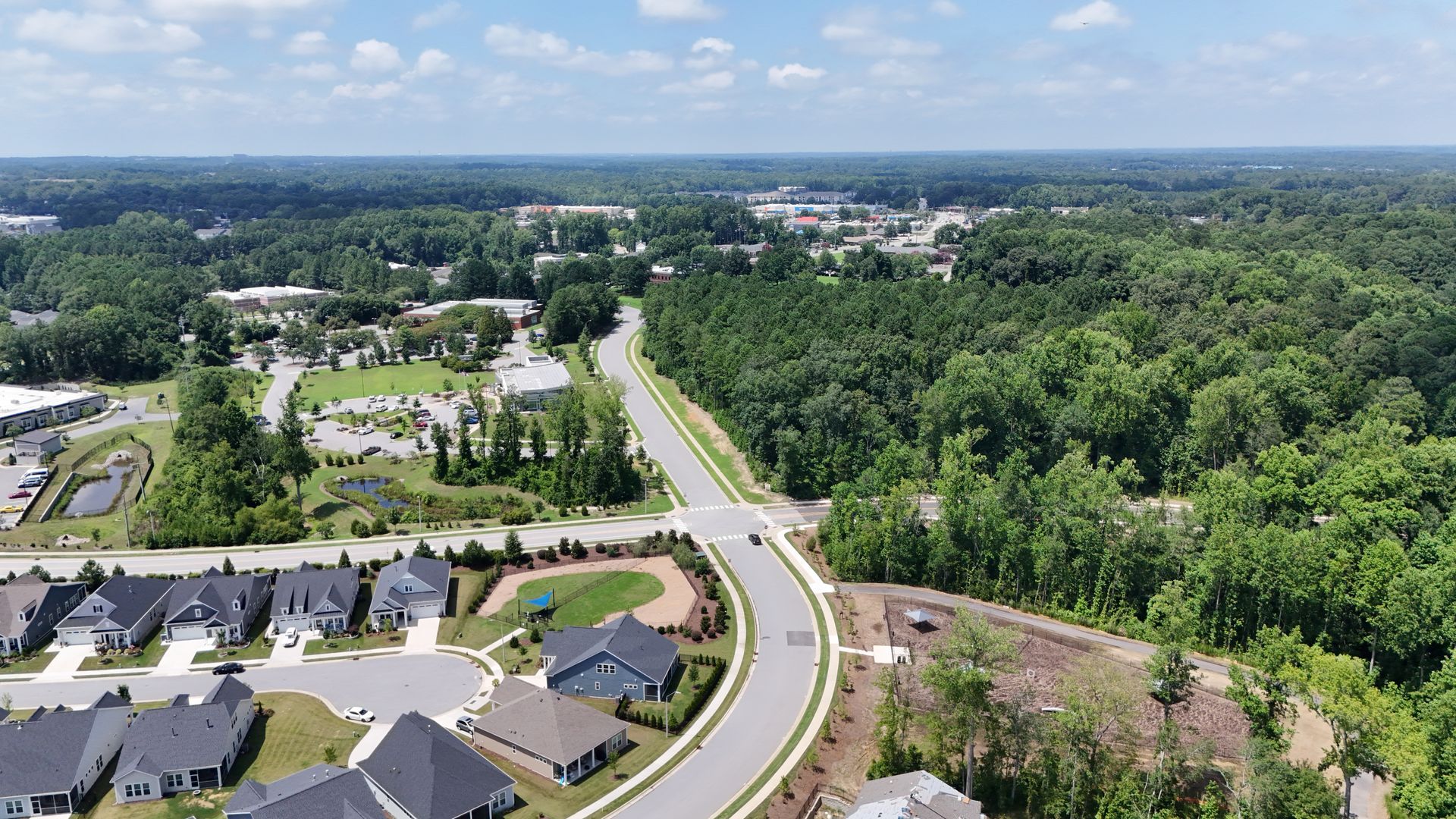 Aerial view of a road curving through a wooded area, with houses and buildings on the left.