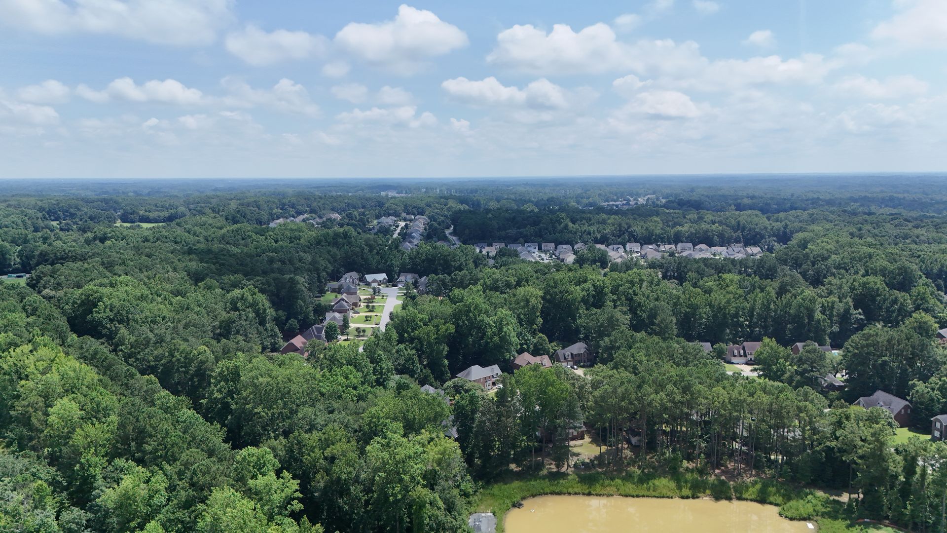 Aerial view of a neighborhood nestled in a lush green forest under a blue sky with fluffy clouds.