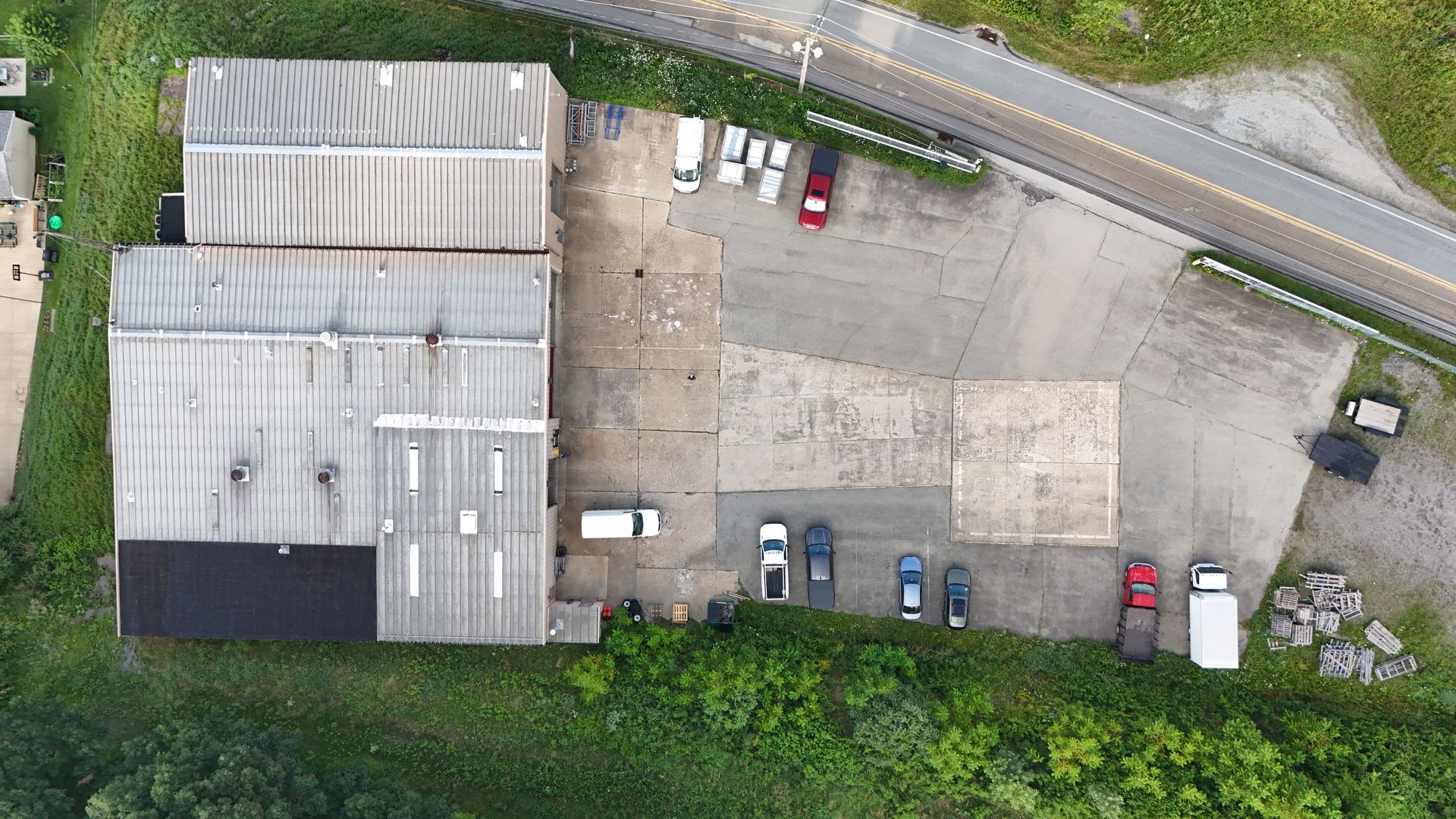 Aerial view of industrial buildings and a parking lot. Cars are parked on concrete. Road and greenery border.