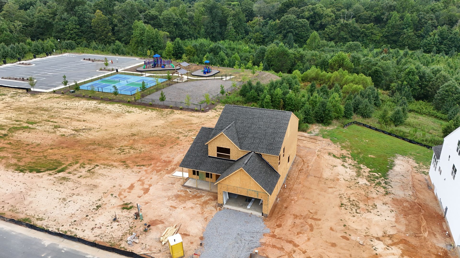 A new house under construction with a dark roof, surrounded by dirt, next to a playground and trees.