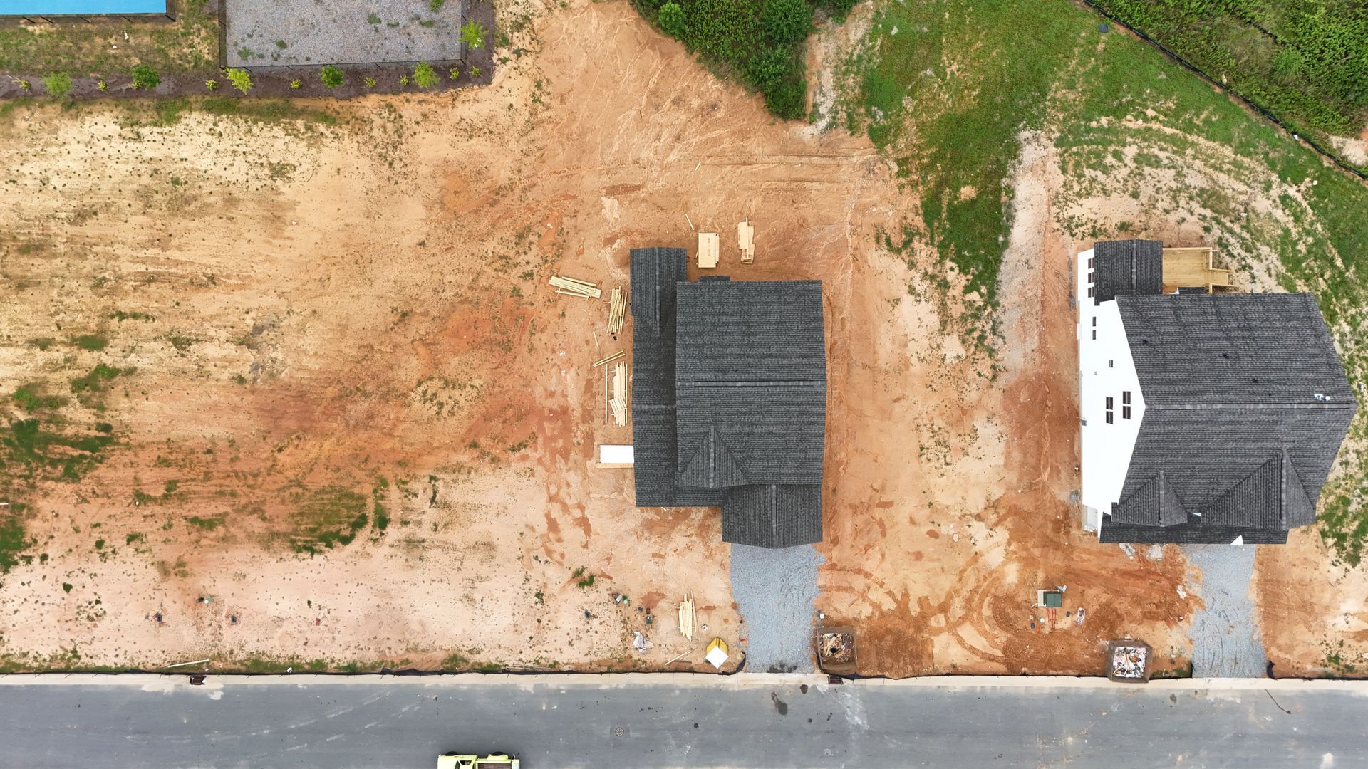 Overhead view of two houses under construction on a dirt lot next to grass and a road.