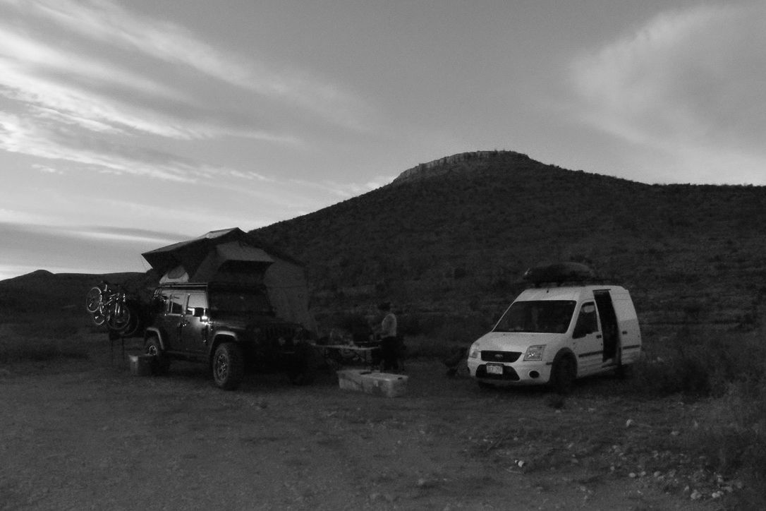 Black and white photo: Two camper vans parked in a desert landscape with a mountain in the background.