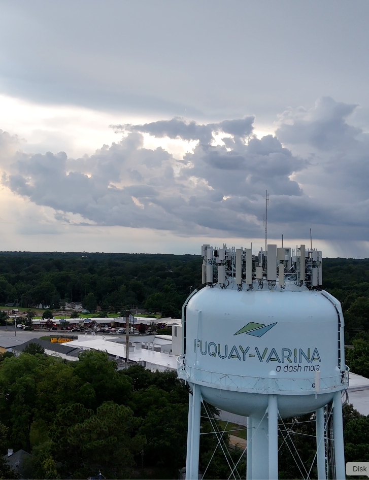 Water tower in Fuquay-Varina, North Carolina, with antennas on top, trees, and a cloudy sky.