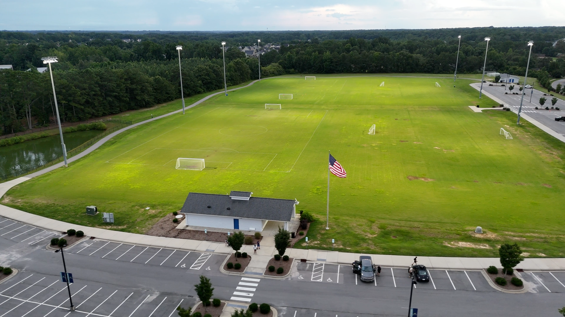 Soccer field with bright green grass, American flag, small building, parking lot, and trees.
