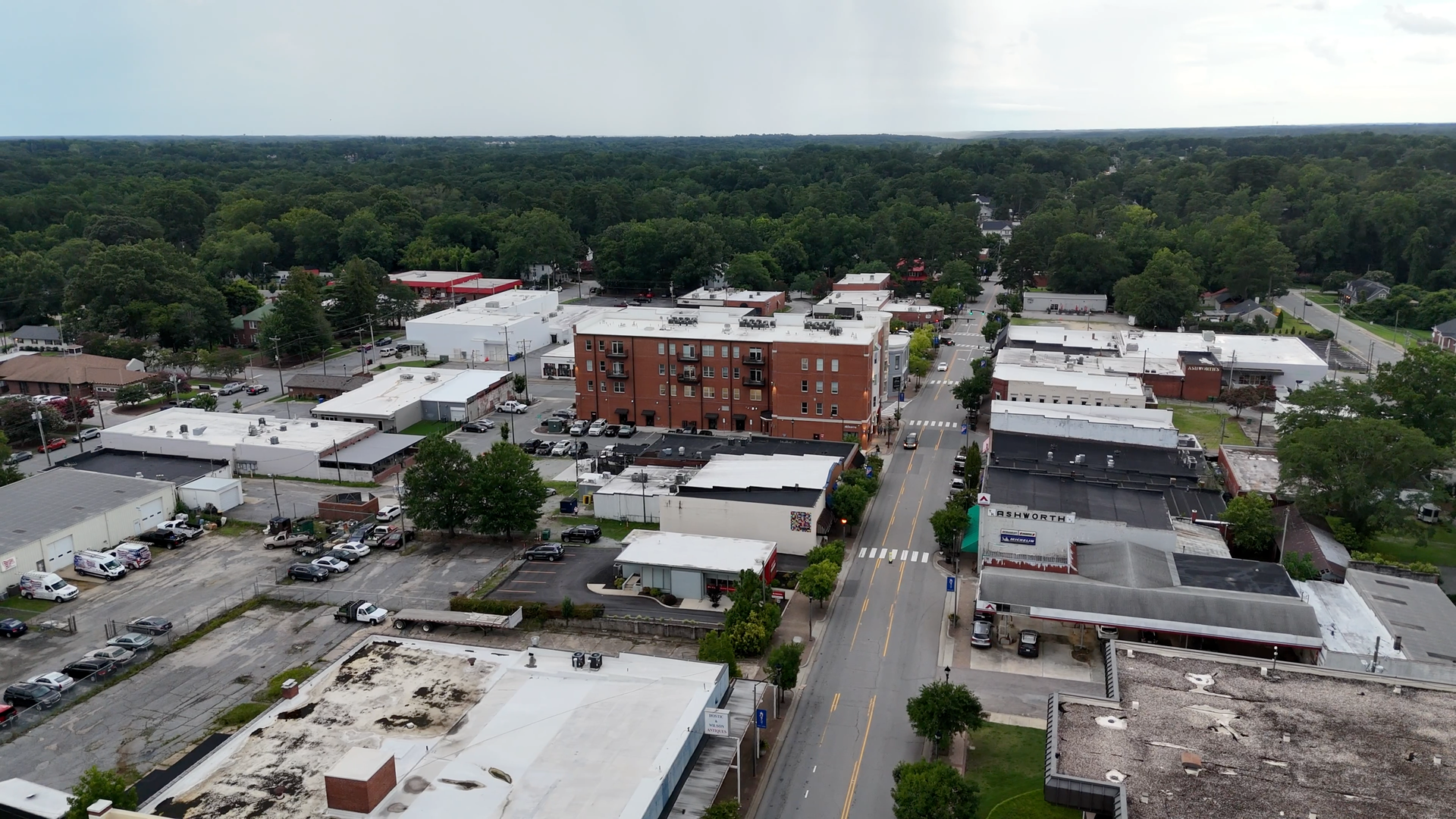 Aerial view of a small town street with shops and a brick building, surrounded by trees under a cloudy sky.
