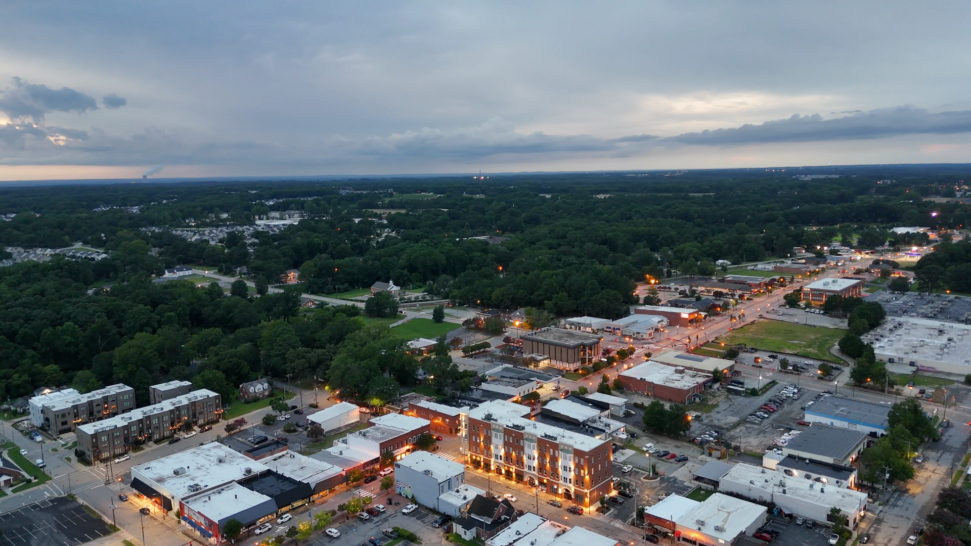 Aerial view of a town at dusk, with lit buildings and a dark green forest backdrop.