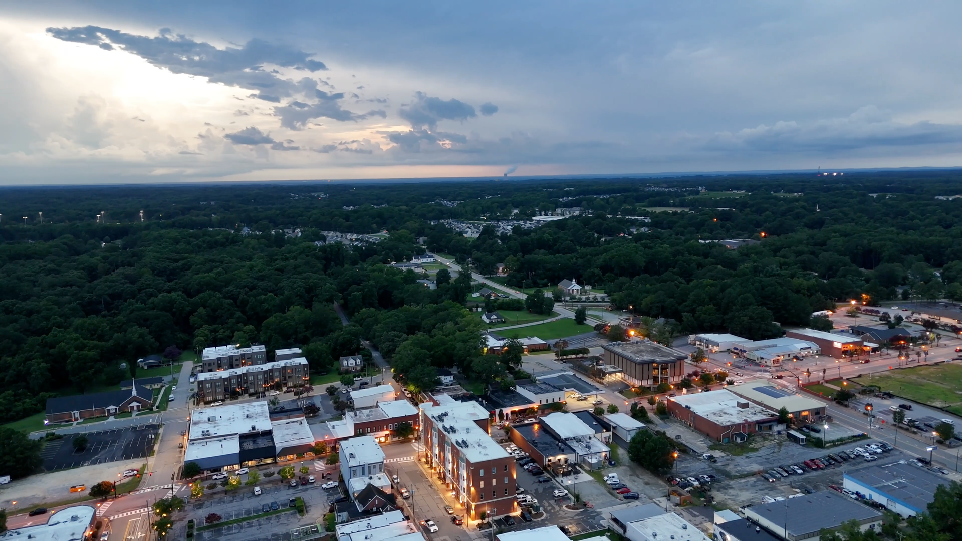 Aerial view of a downtown area at dusk, with lit buildings and surrounding dark trees under a cloudy sky.