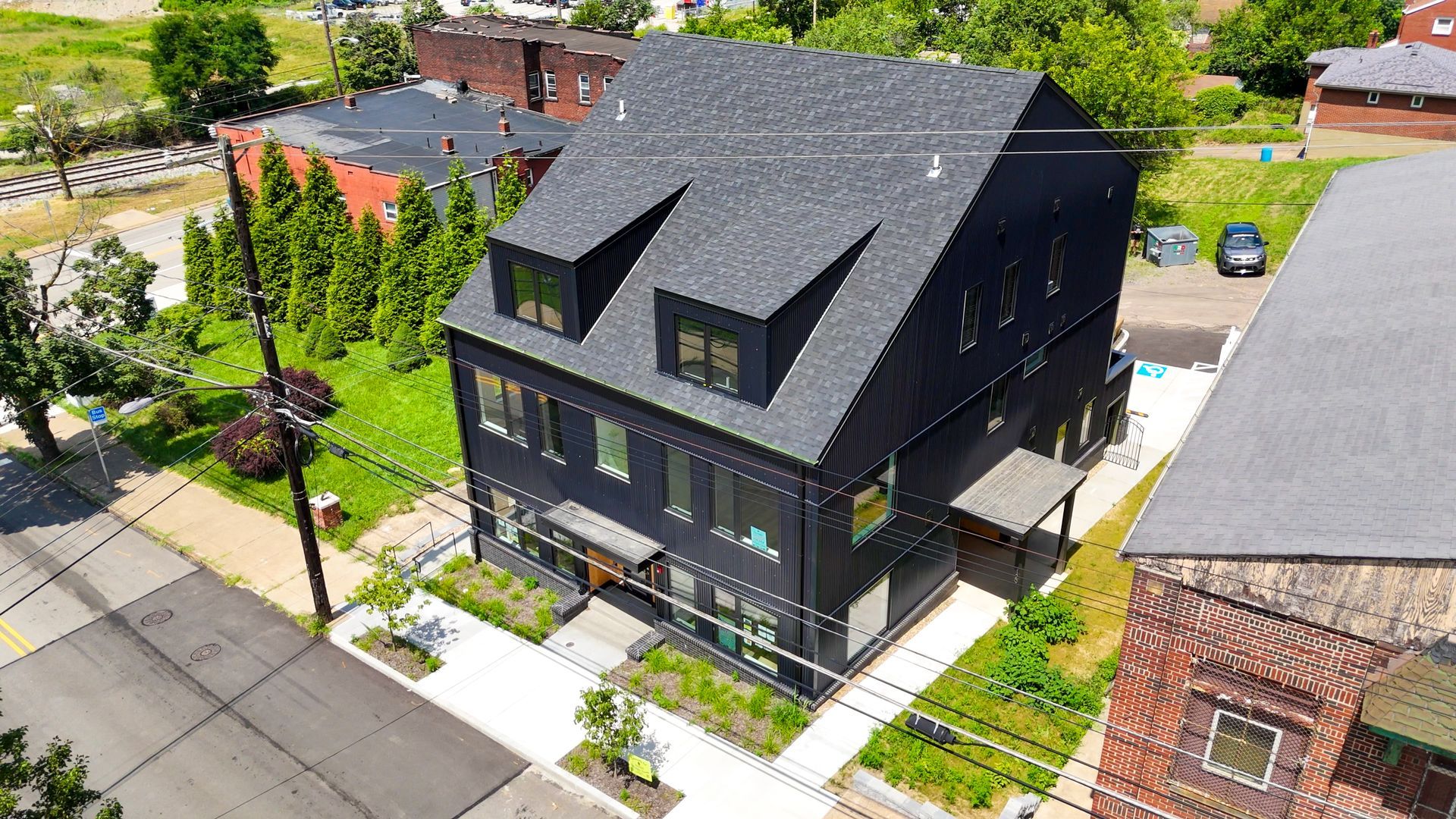 Black modern multi-story building with dark roof, dormers, and landscaping. Street view, sunny day.