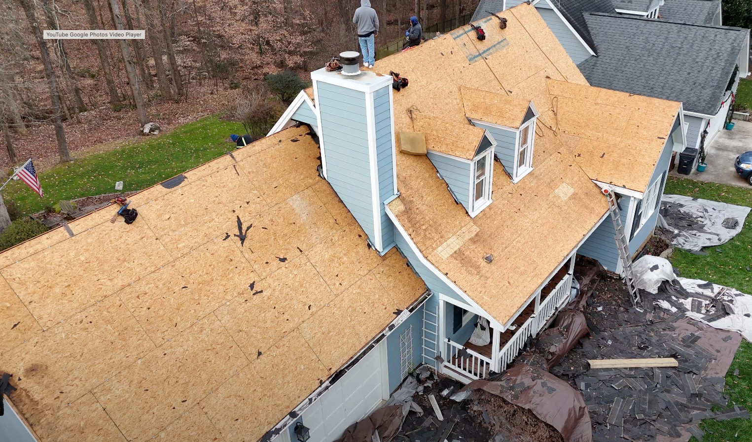 House with damaged roof, being repaired. Light brown shingles, blue siding. Workers on roof.