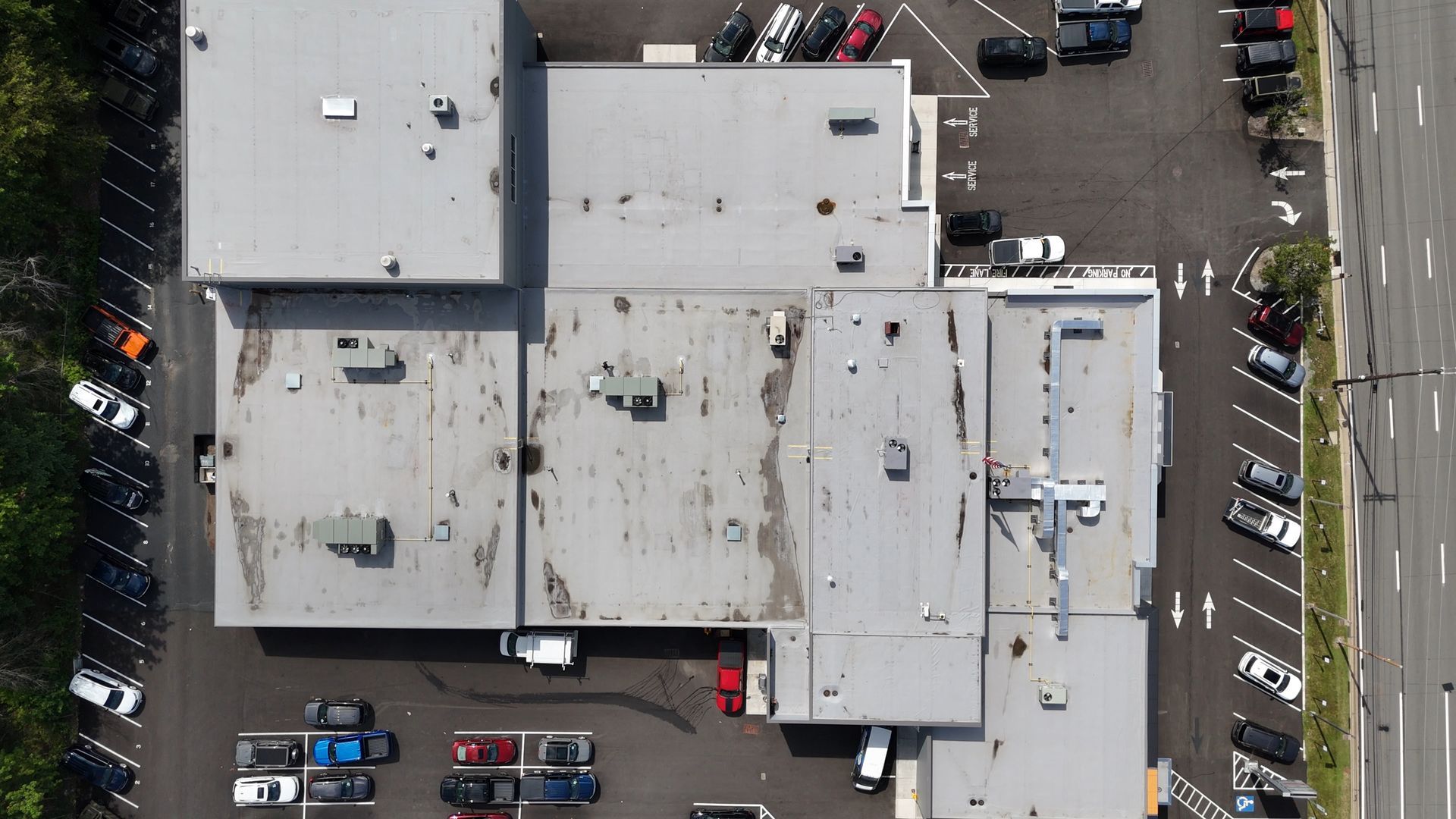 Aerial view of a commercial building with multiple flat gray roofs, surrounded by parking lots with cars.