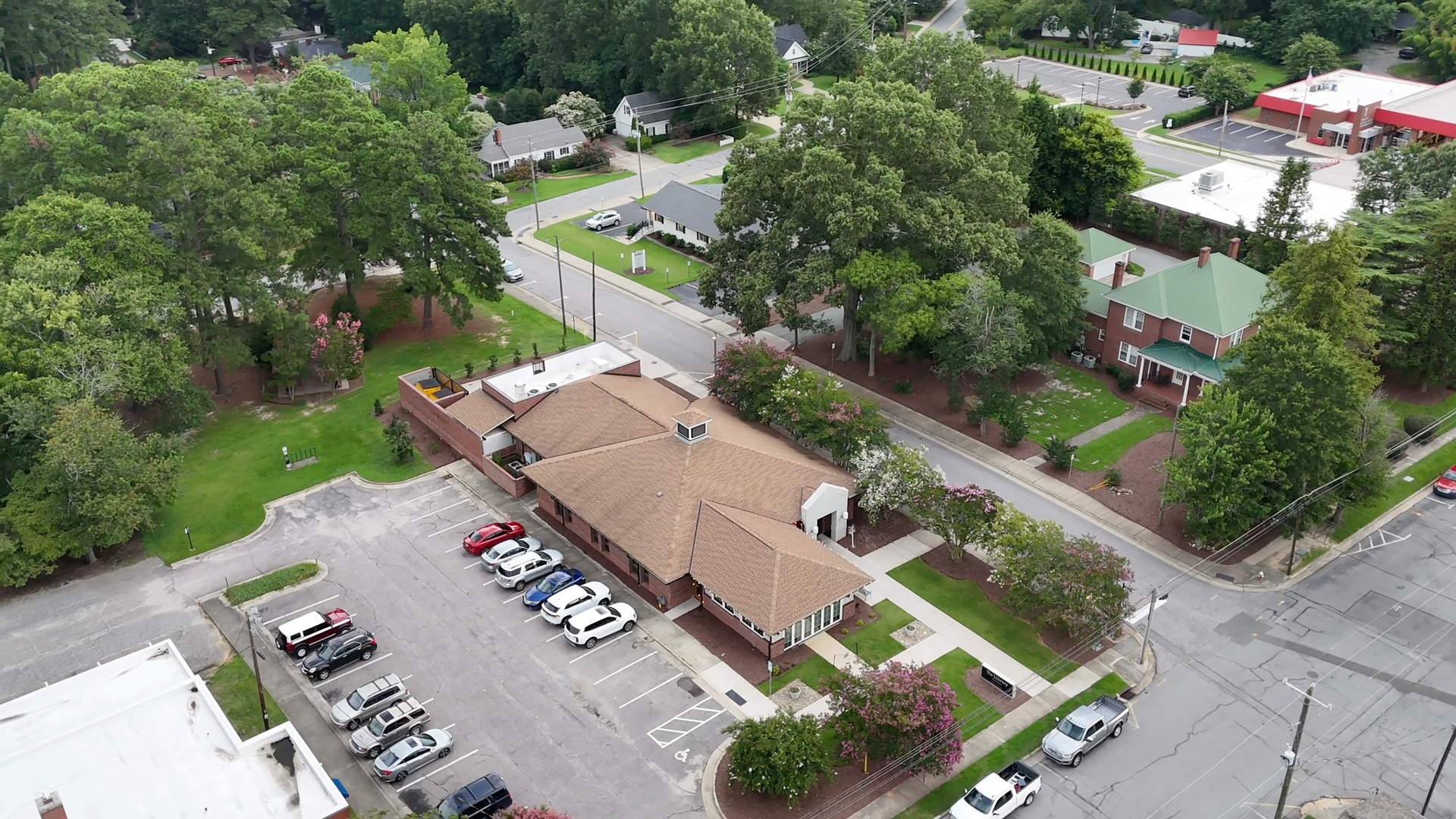 Aerial view of a brown-roofed building with cars parked in front; surrounded by green trees and a street.